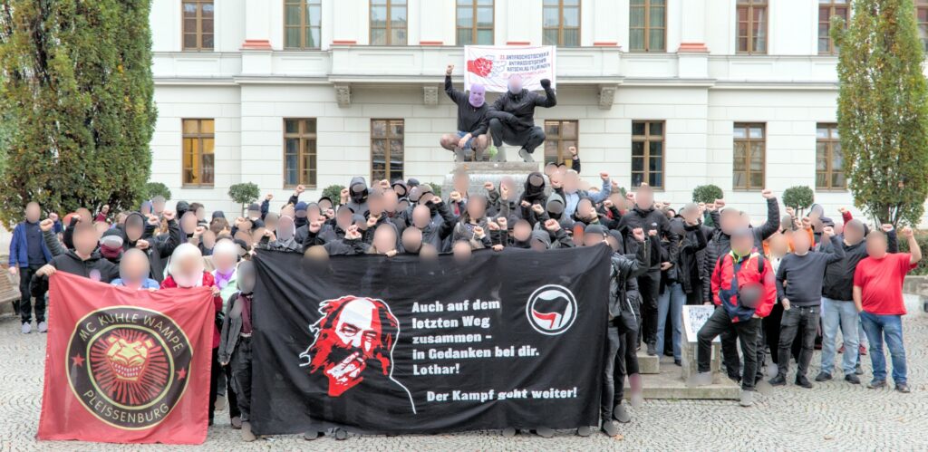 Eine Reihe von Menschen steht vor dem mon ami in Weimar, einige strecken die Faust nach oben. Es sind 2 Transparente zu sehen, auf dem einen steht "MC KUHLE WAMPE - PLEISSENBURG" auf dem anderen ist eine Illustration von Lothar König und es steht "Auch auf dem letzten Weg zusammen - in Gedanken bei dir. Lothar! Der Kampf geht weiter!"